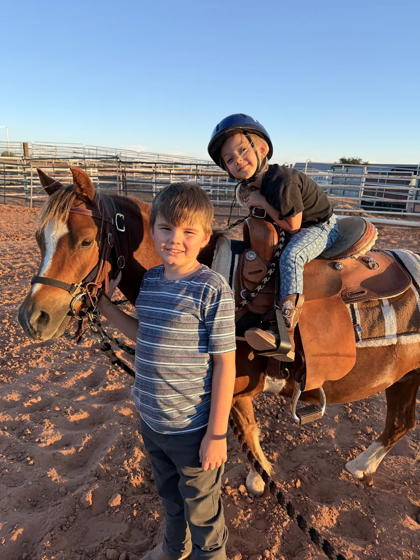 Two kids with a saddled pony in the arena, one in a riding helmet